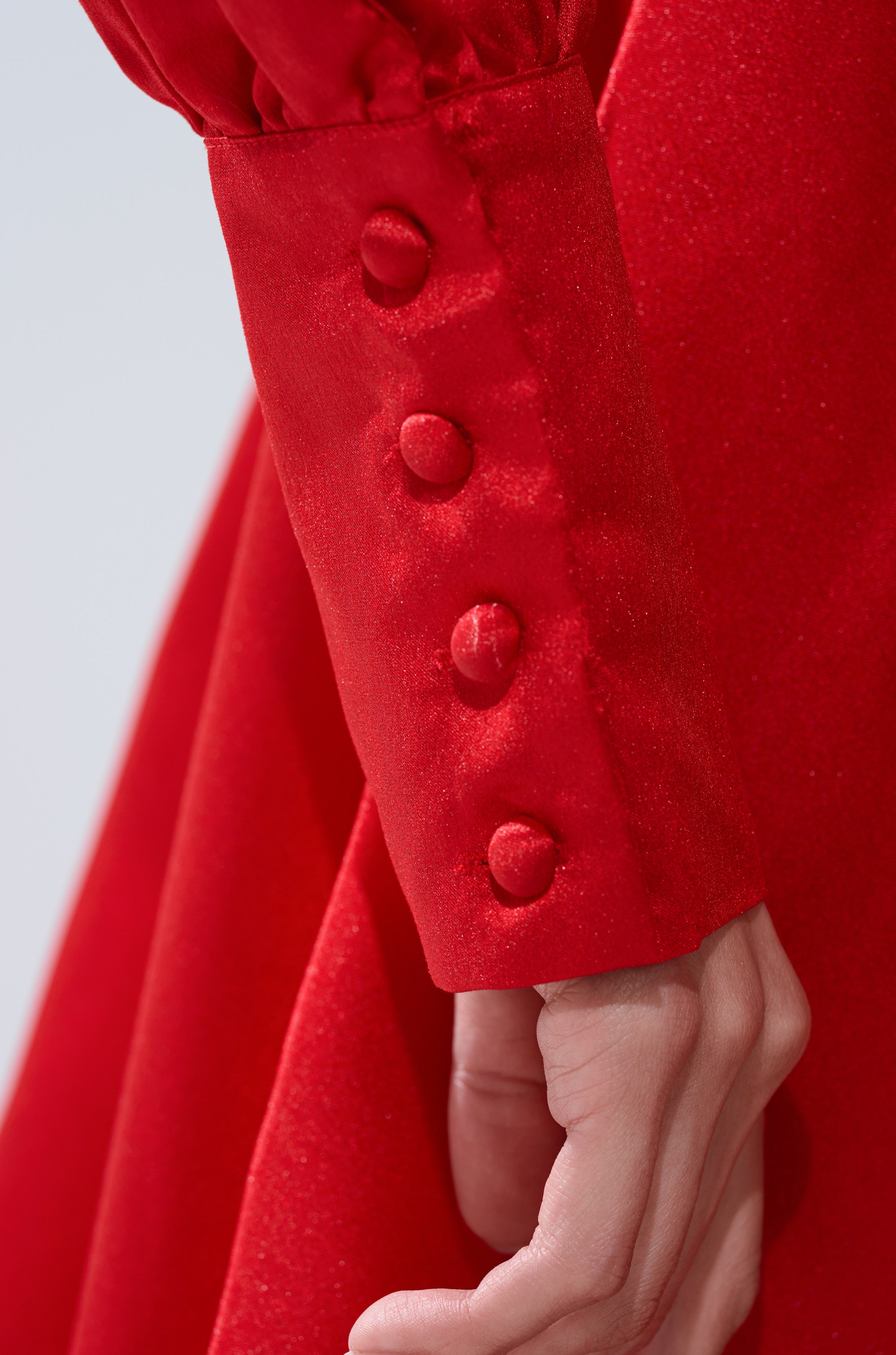 A close-up of a hand wearing the ELOUISE HI LOW DRESS IN RED, featuring fabric-covered cuff buttons. The dress’s soft, slightly shiny texture adds an elegant touch.