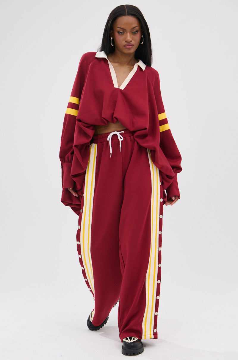 A woman wears the GAME OVER RELAXED PANT—a loose-fitting, deep red style with yellow and white stripes, a white drawstring waist, and black shoes—creating a sporty-chic look against a plain light background.