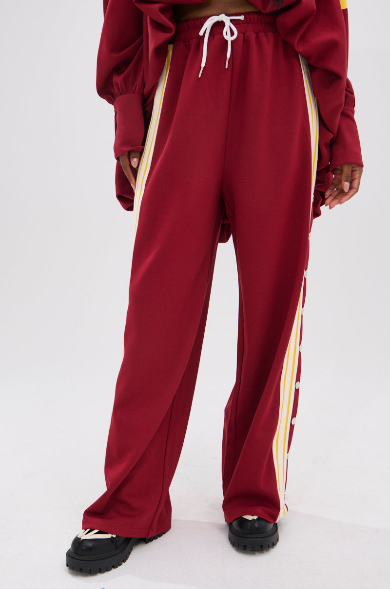 Only the lower half is visible: loose-fitting maroon GAME OVER RELAXED PANT with yellow and white side stripes, paired with black chunky shoes and a matching jacket for a 90’s-inspired sporty-chic look.