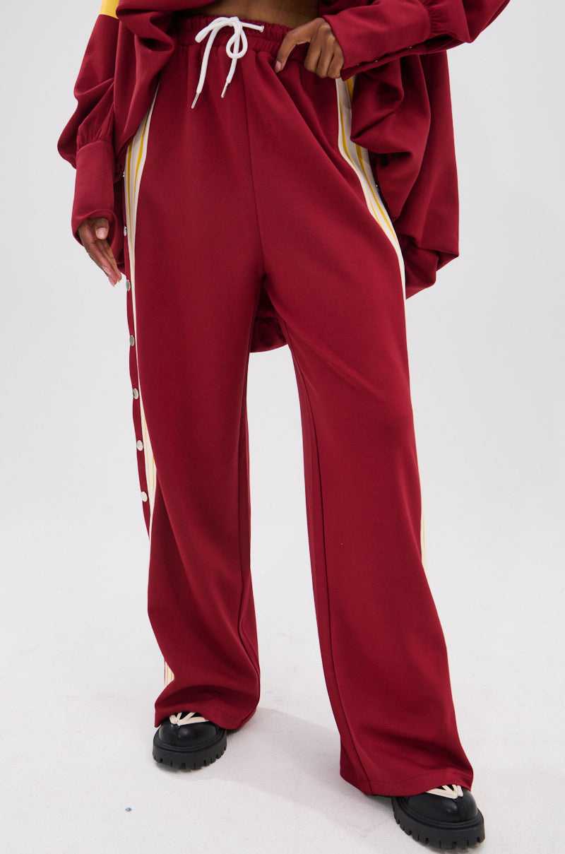 A person wears loose-fitting maroon GAME OVER RELAXED PANT with white and yellow side stripes, paired with a matching oversized jacket and black shoes, standing against a white background for a sporty-chic look.