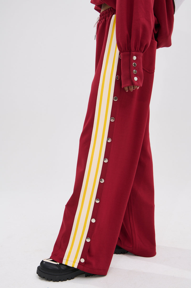 Someone wears the red GAME OVER RELAXED PANT, featuring yellow and white side stripes and silver snap buttons, paired with a matching top and chunky black shoes for a sporty-chic style against a plain light background.