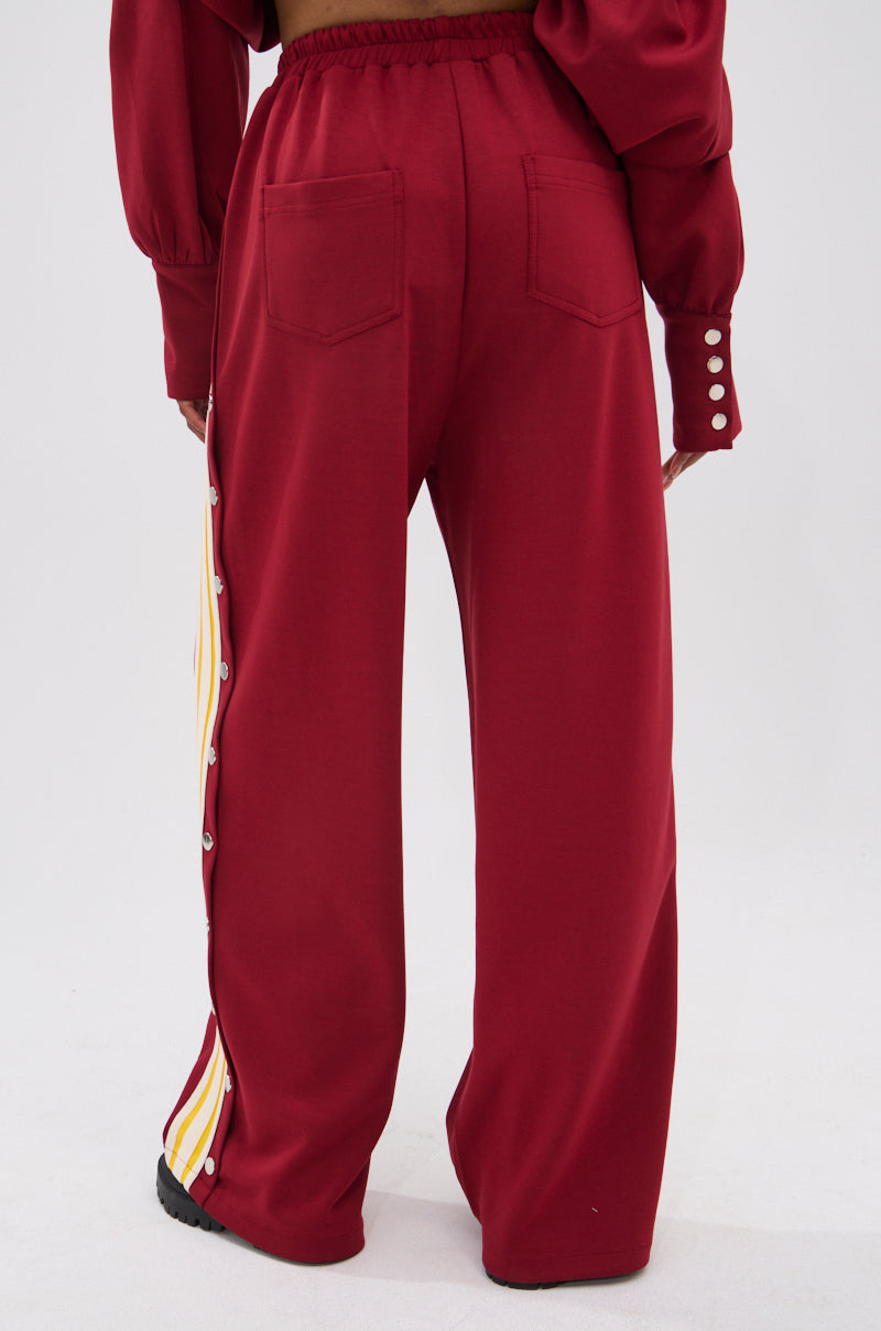 A person is seen from behind wearing the GAME OVER RELAXED PANT—loose-fit with yellow and white athletic stripes, silver snap buttons down the sides, back patch pockets—paired with a matching red top.