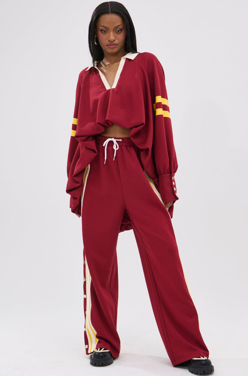 A woman exudes confidence in GAME OVER RELAXED PANT, featuring a loose maroon fit with yellow stripes and a cropped top for a sporty-chic vibe. Black chunky shoes complete her 90s-inspired look.