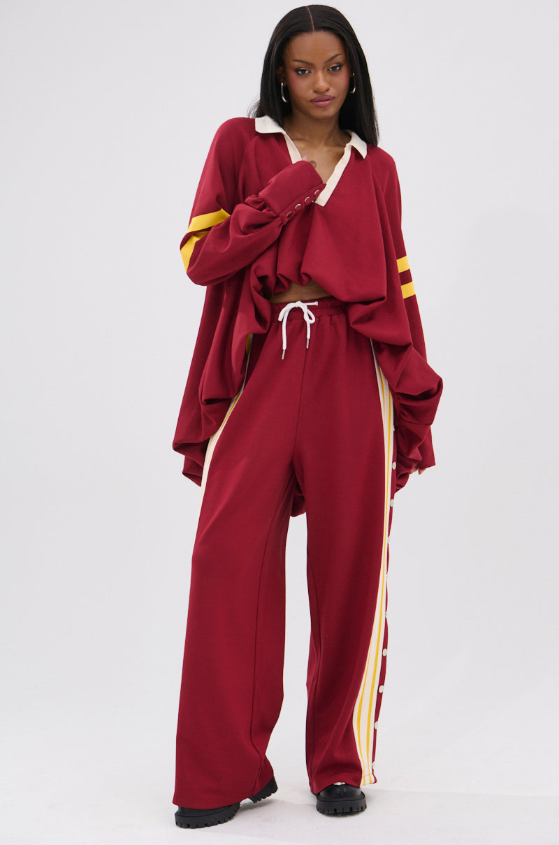 A woman poses confidently in the GAME OVER RELAXED PANT, a loose-fitting, burgundy tracksuit with yellow stripes and 90s-inspired style. She pairs it with a white-collared top, showing her midriff, and finishes the sporty-chic look with black shoes.