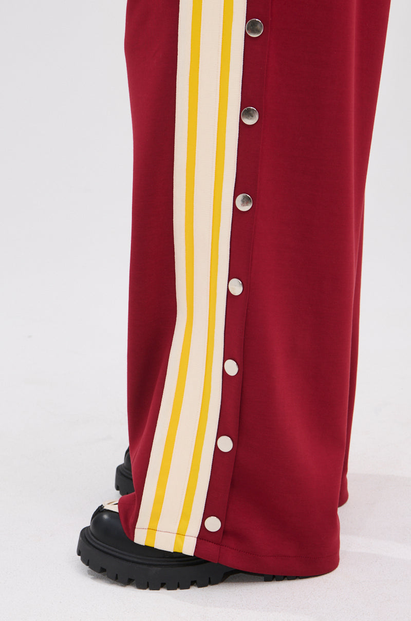 A person wears maroon GAME OVER RELAXED PANT with yellow and white side stripes and silver snap buttons, styled with black chunky shoes for a sporty-chic look.