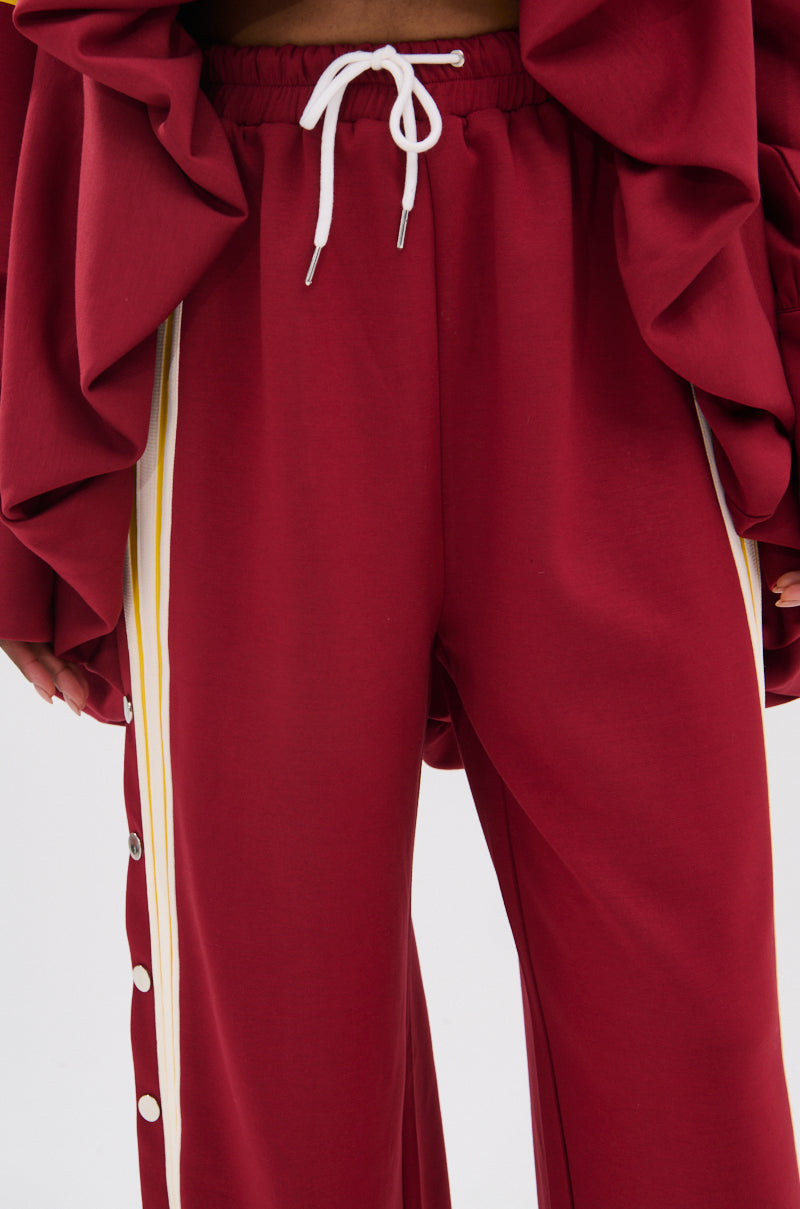 Wearing the GAME OVER RELAXED PANT, a 90s-inspired style with a white drawstring, yellow and white side stripes, snap buttons, and a sporty-chic finish. Maroon ruffled sleeves on top add extra flair.