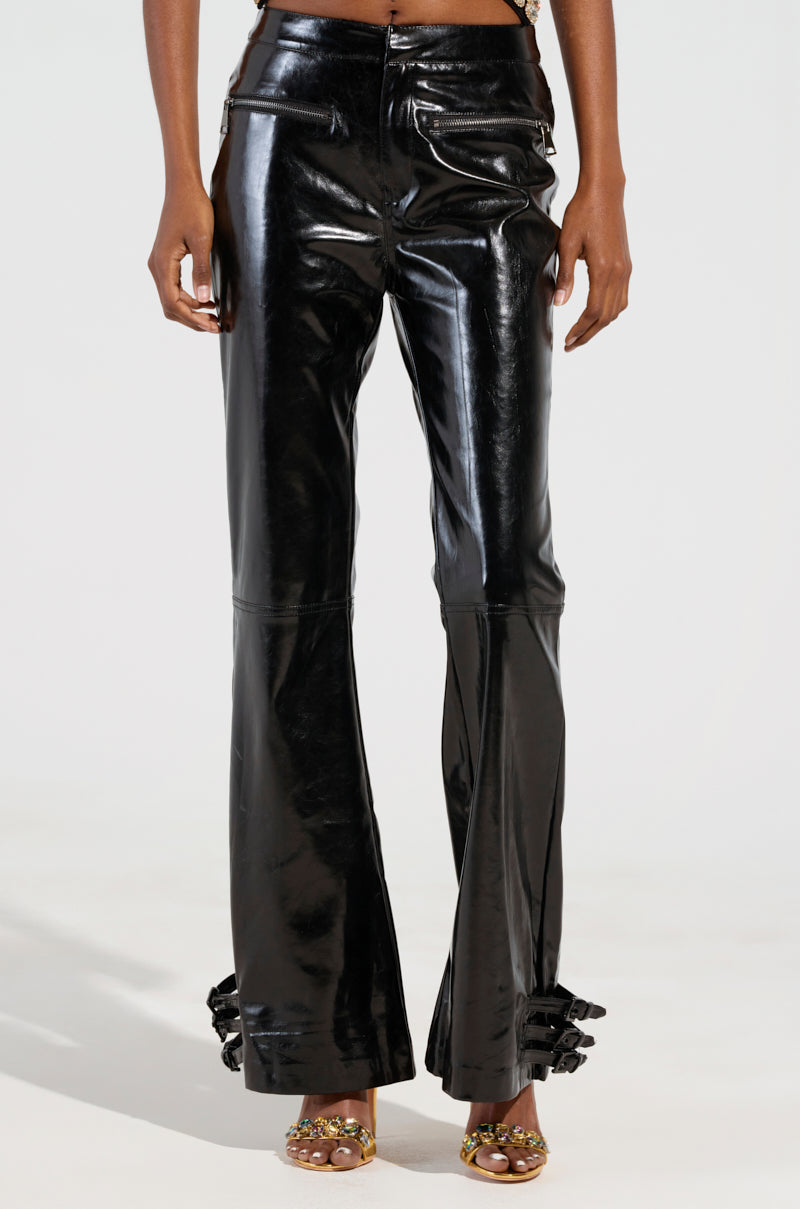 A person wearing the FEELS SO GOOD TROUSER, made of glossy faux patent leather with zippers and flared pantlegs featuring decorative bows near the ankles, is paired with open-toe sandals adorned with floral embellishments against a white background.