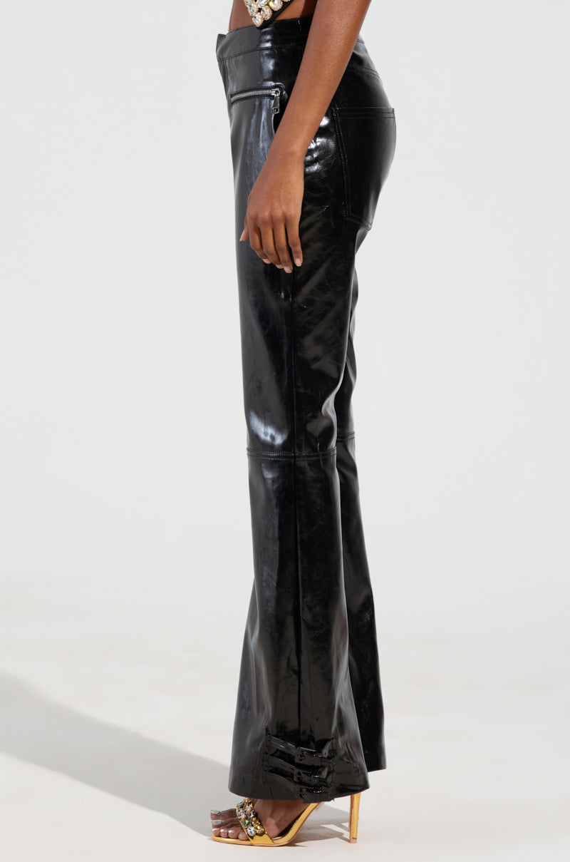 Wearing the FEELS SO GOOD TROUSER, a person stands sideways against a plain backdrop, highlighting the seam details of these glossy faux patent leather trousers. These fitted pants feature flared legs and are perfectly paired with heeled sandals.