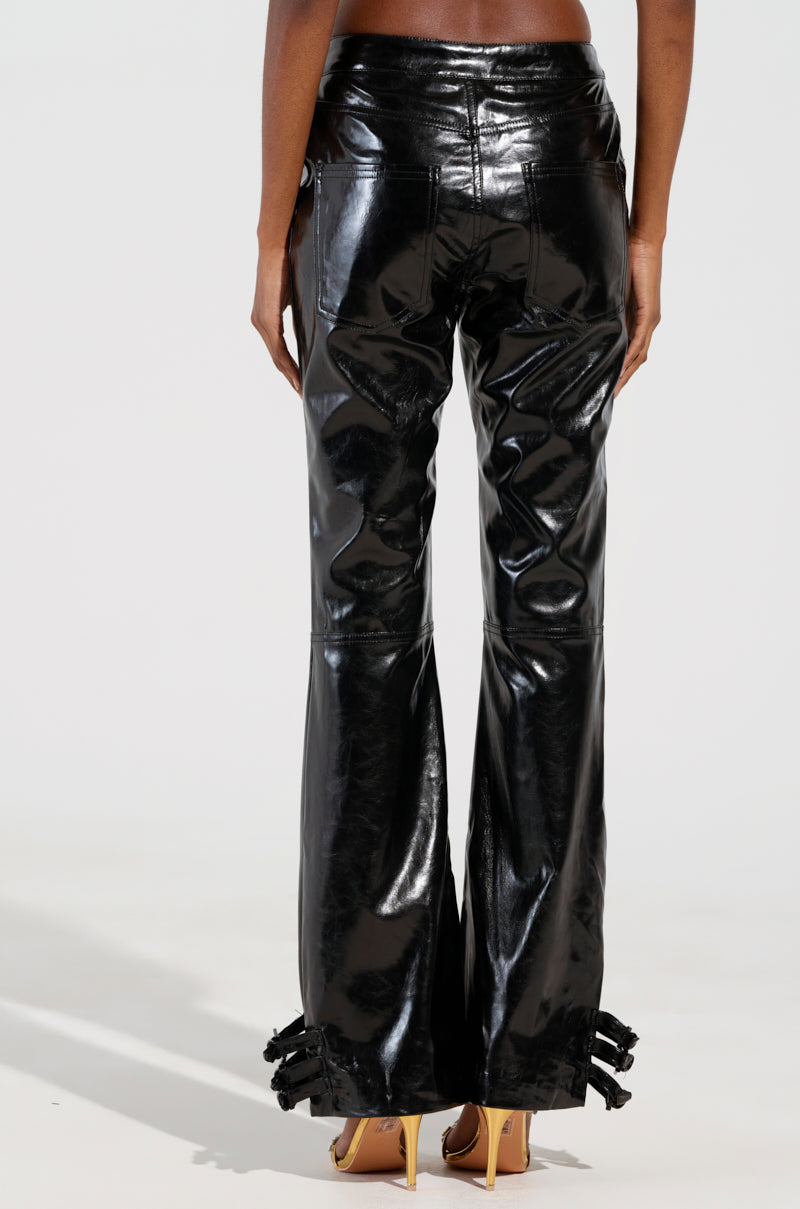 A person wearing the "FEELS SO GOOD TROUSER" in shiny black faux patent leather, featuring a bell-bottom design and black tassels at the hem, stands with their back to the camera. These mid-rise fit trousers, with visible seams, are paired with tan high-heeled shoes.