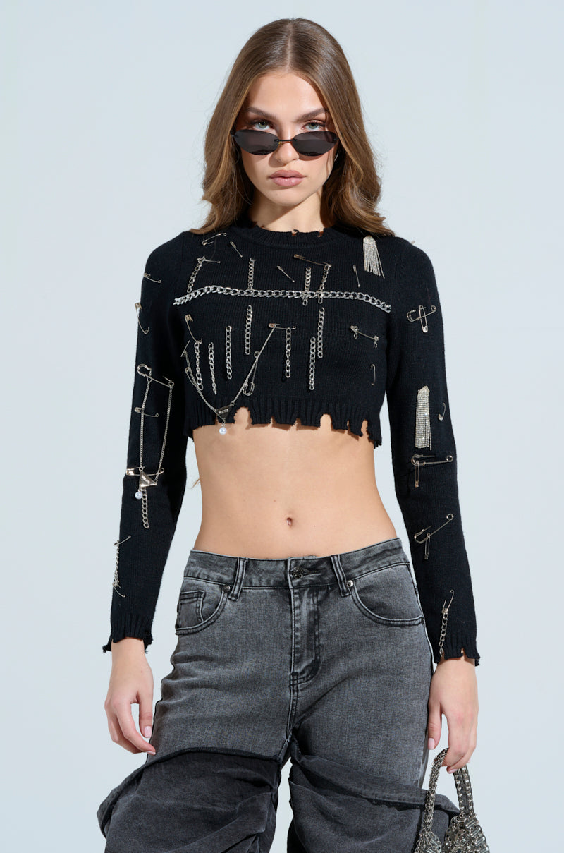 Sporting a grunge-chic look, an individual with long hair wears sunglasses and the DEJA EMBELLISHED CROP SWEATER, featuring metallic safety pins. They team it with high-waisted, two-tone jeans with a frayed hem and carry a textured handbag against a plain background.
