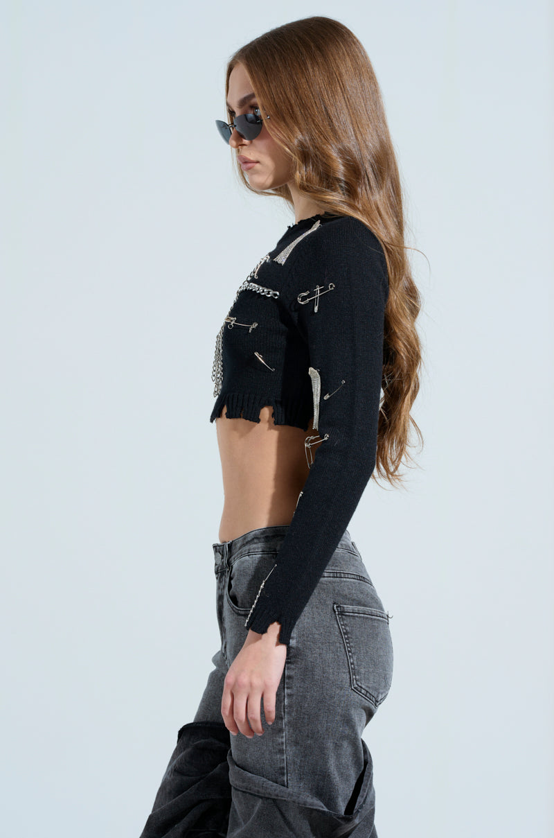 A person with long brown hair, sunglasses, and a grunge-chic DEJA EMBELLISHED CROP SWEATER with metallic safety pins stands in profile against a plain background, paired with gray jeans.