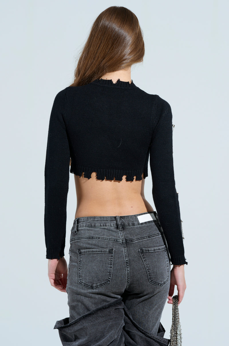 With grunge-chic flair, a long-haired person wears the DEJA EMBELLISHED CROP SWEATER—black, long-sleeved, and adorned with metallic safety pins—paired with high-waisted gray jeans. Facing away, they hold a small handbag against a plain backdrop.