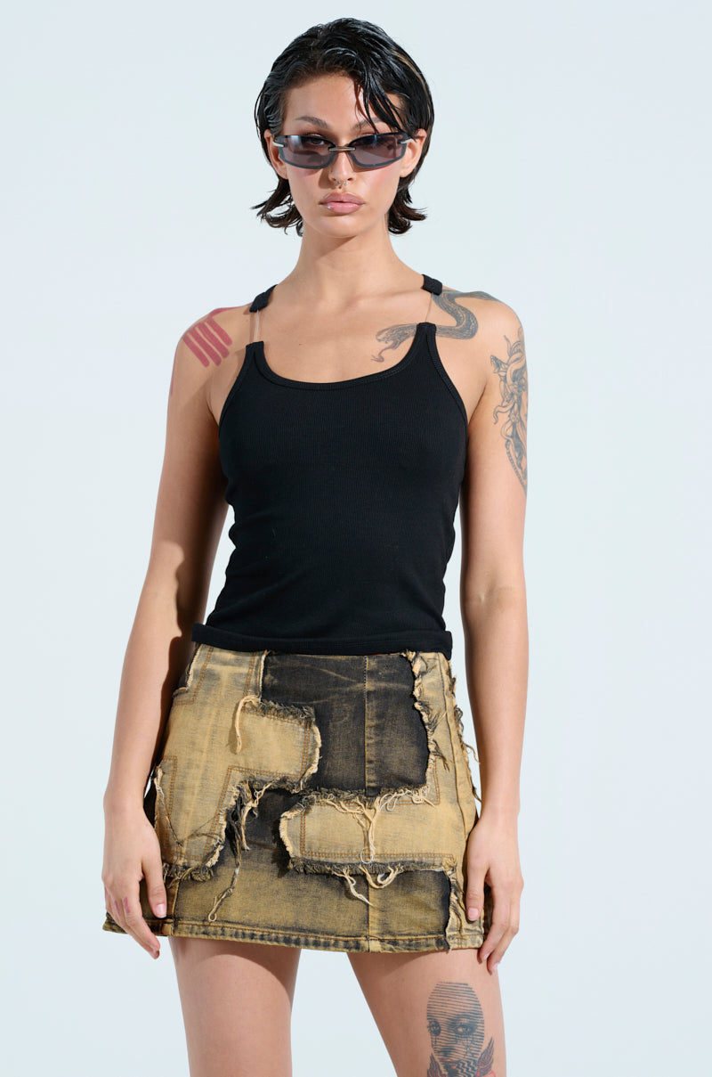 A person wearing sunglasses, the BAD HABIT STRAPPY TANK TOP IN BLACK (bodycon fit), and a distressed denim skirt with patchwork. They have short dark hair and arm tattoos, standing against a plain light background.