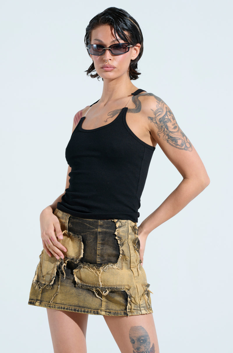A person with short dark hair stands confidently, wearing sunglasses and the BAD HABIT STRAPPY TANK TOP IN BLACK paired with a distressed denim skirt. Tattoos adorn their arms and legs against a plain background.