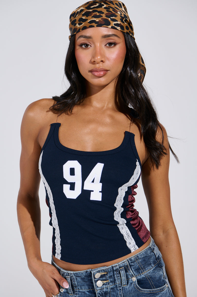 In the JESSICA RAZER BACK TOP, a dark blue sporty-chic tank featuring the number "94" and lace accents, one exudes confidence. Paired with denim bottoms and elegantly styled with long, dark hair wrapped in a leopard print headscarf, this look embodies effortless AKIRA Label style.