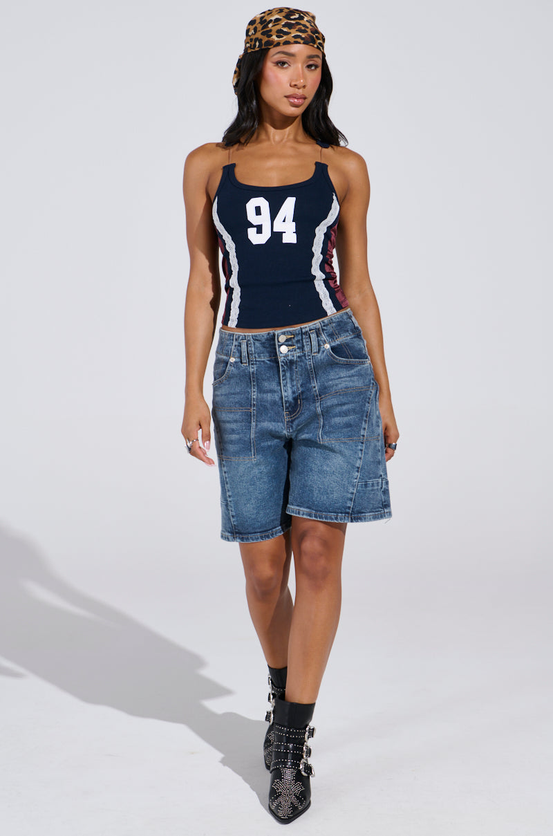 Wearing the sporty-chic JESSICA RAZER BACK TOP in navy, numbered "94," paired with denim shorts and snakeskin ankle boots, a person stands confidently. A leopard print headscarf complements their long hair against a plain white background, showcasing AKIRA Label's street-savvy style.