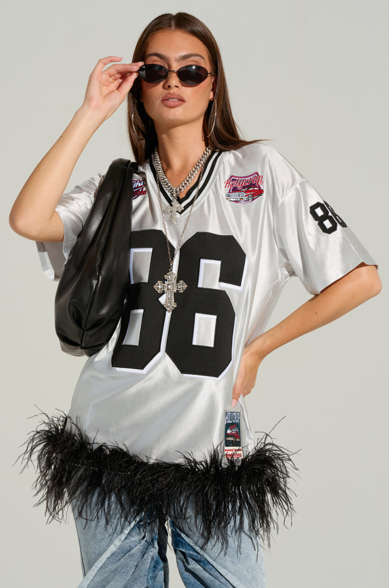 A person wearing the GO SPORTS FEATHER TRIM JERSEY DRESS in silver, featuring the number 86 and sports-inspired embroidered graphics, poses confidently. The dress boasts an oversized silhouette with patches and black feather trim at the hem. They're accessorized with large sunglasses, layered necklaces, a black shoulder bag, and light blue jeans.