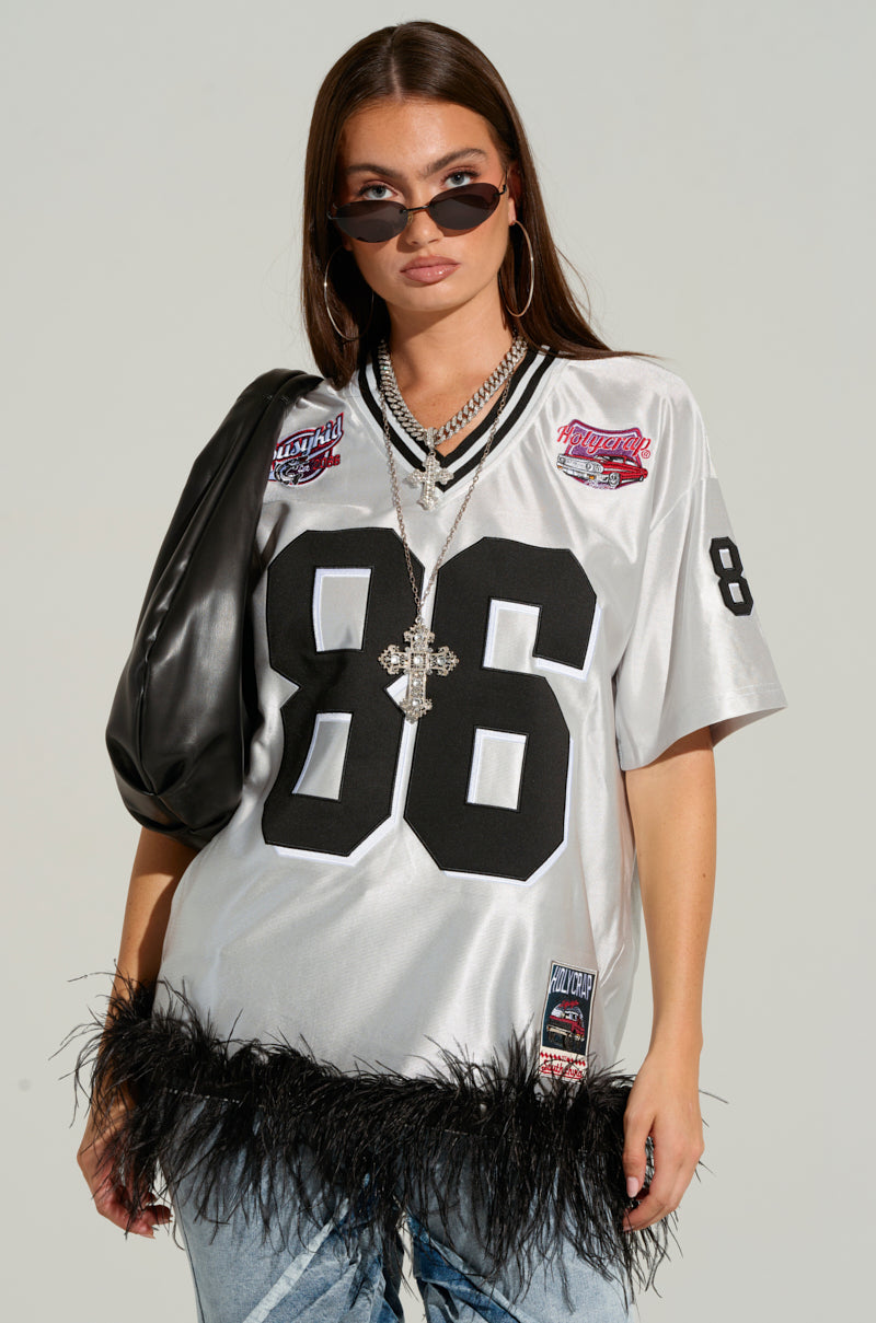 A woman with long brown hair wears a GO SPORTS FEATHER TRIM JERSEY DRESS, featuring sports-inspired embroidered graphics and the number 86. She accessorizes with black sunglasses, hoop earrings, layered necklaces, and carries a black bag on her shoulder.