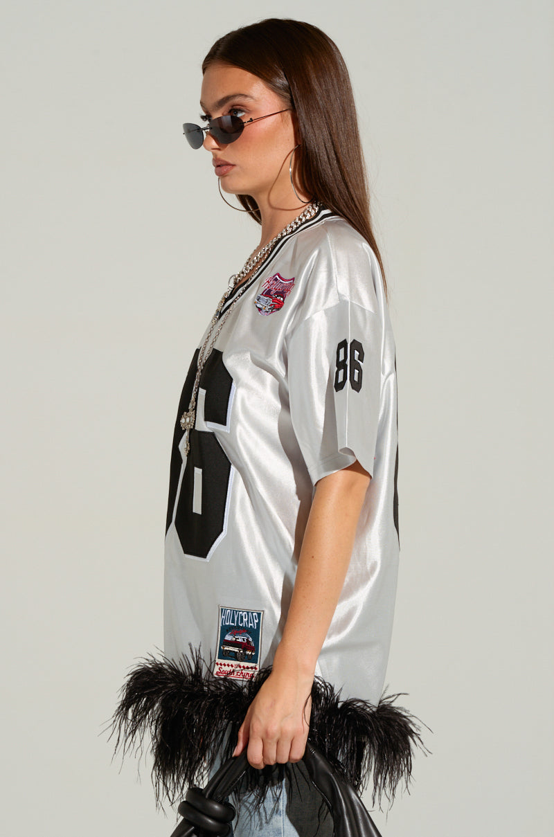 A woman with long brown hair wears sunglasses and the GO SPORTS FEATHER TRIM JERSEY DRESS, featuring black stripes and the number 86. Complemented by a statement necklace, she carries a purse, showcasing an oversized silhouette highlighted by sports-inspired embroidered graphics, all set against a plain grey background.