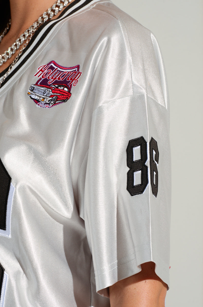 A close-up of a person wearing the GO SPORTS FEATHER TRIM JERSEY DRESS, an oversized silver sports jersey with black trim. There's a black number "86" on the sleeve and sports-inspired embroidered graphics with the word "Hollywood" on the chest. The person also wears a gold chain necklace.
