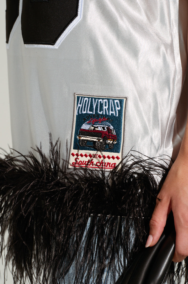 Close-up of a person wearing the GO SPORTS FEATHER TRIM JERSEY DRESS, featuring an oversized silhouette with a patch that reads "HOLYCRAP" and sports-inspired embroidered graphics of a car alongside the text "South Lima." The dress is elegantly accented with black feather-like trim, while the person holds a black object on their right.
