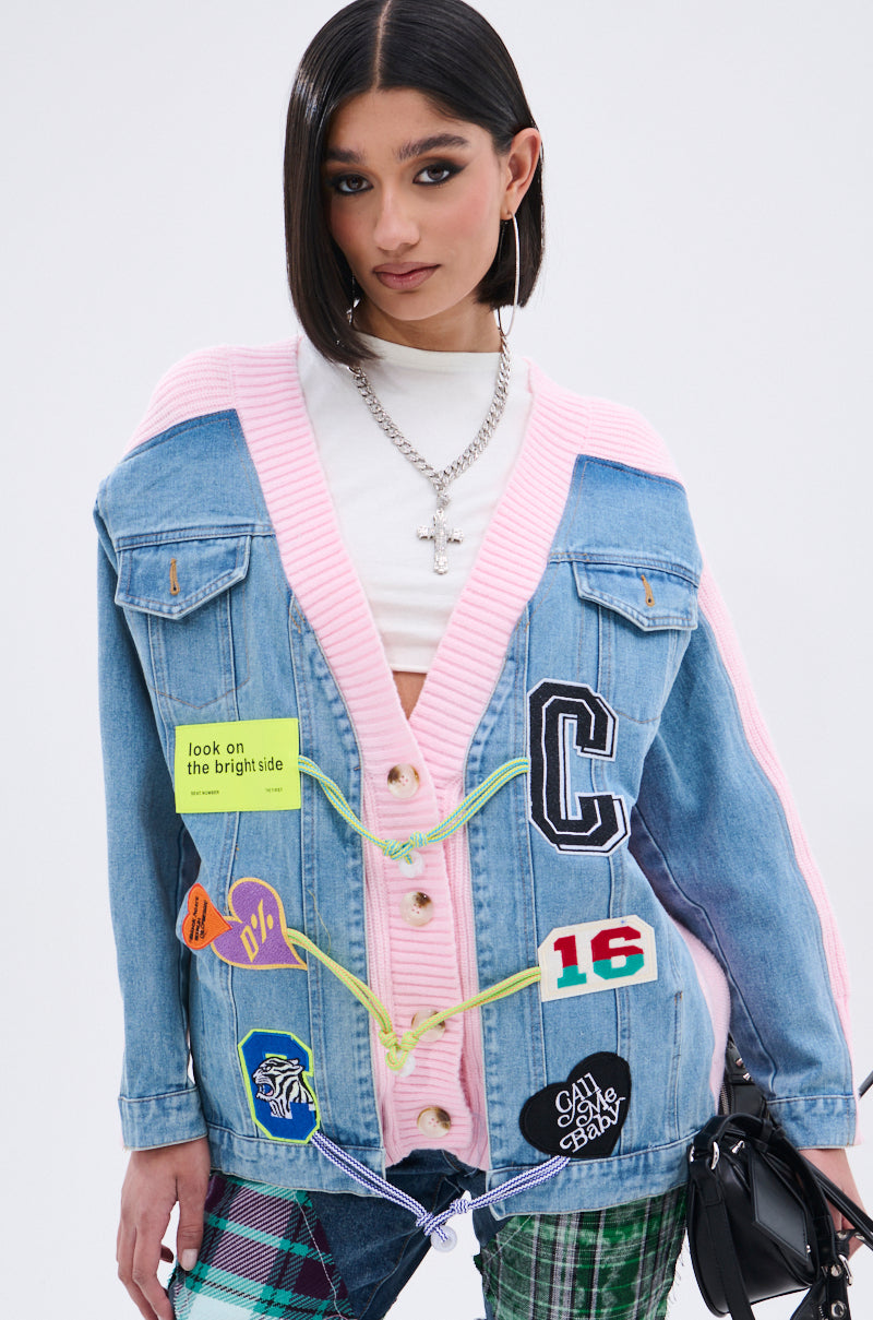 A woman with straight dark hair wears a pink and blue denim SURGICAL SPLICED CARDIGAN with colorful patches and cords over a white turtleneck, paired with green plaid pants, silver jewelry, and holding a black bag. 0