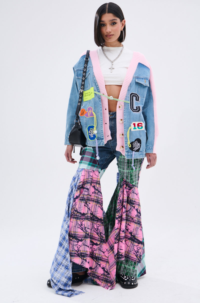 A woman in a cropped white top, SURGICAL SPLICED CARDIGAN denim jacket, and flared patchwork jeans with blue, pink, and green patterns holds a black purse against a plain white background. 9