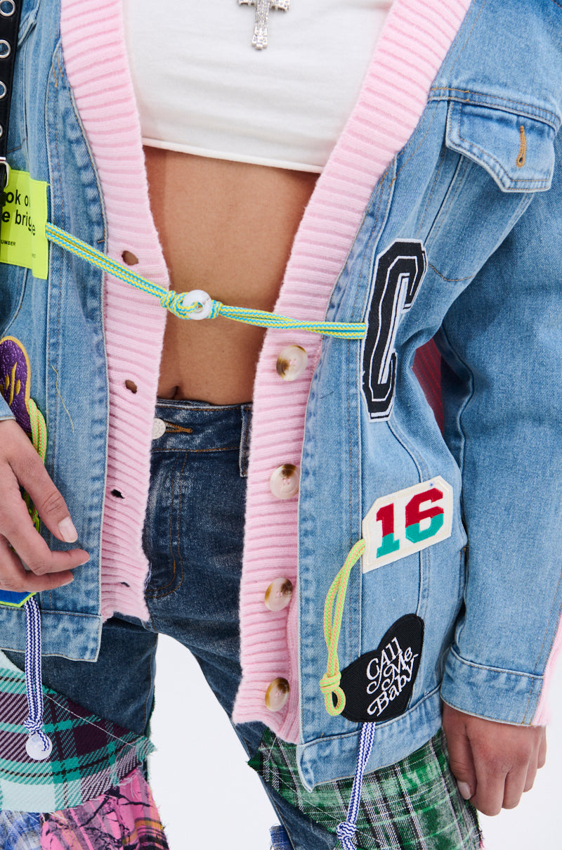 A person wears the SURGICAL SPLICED CARDIGAN, a layered denim and pink cardigan with colorful patches, graphic buttons, and drawstrings, paired with plaid and denim pants; only the torso and part of the legs are visible. 11