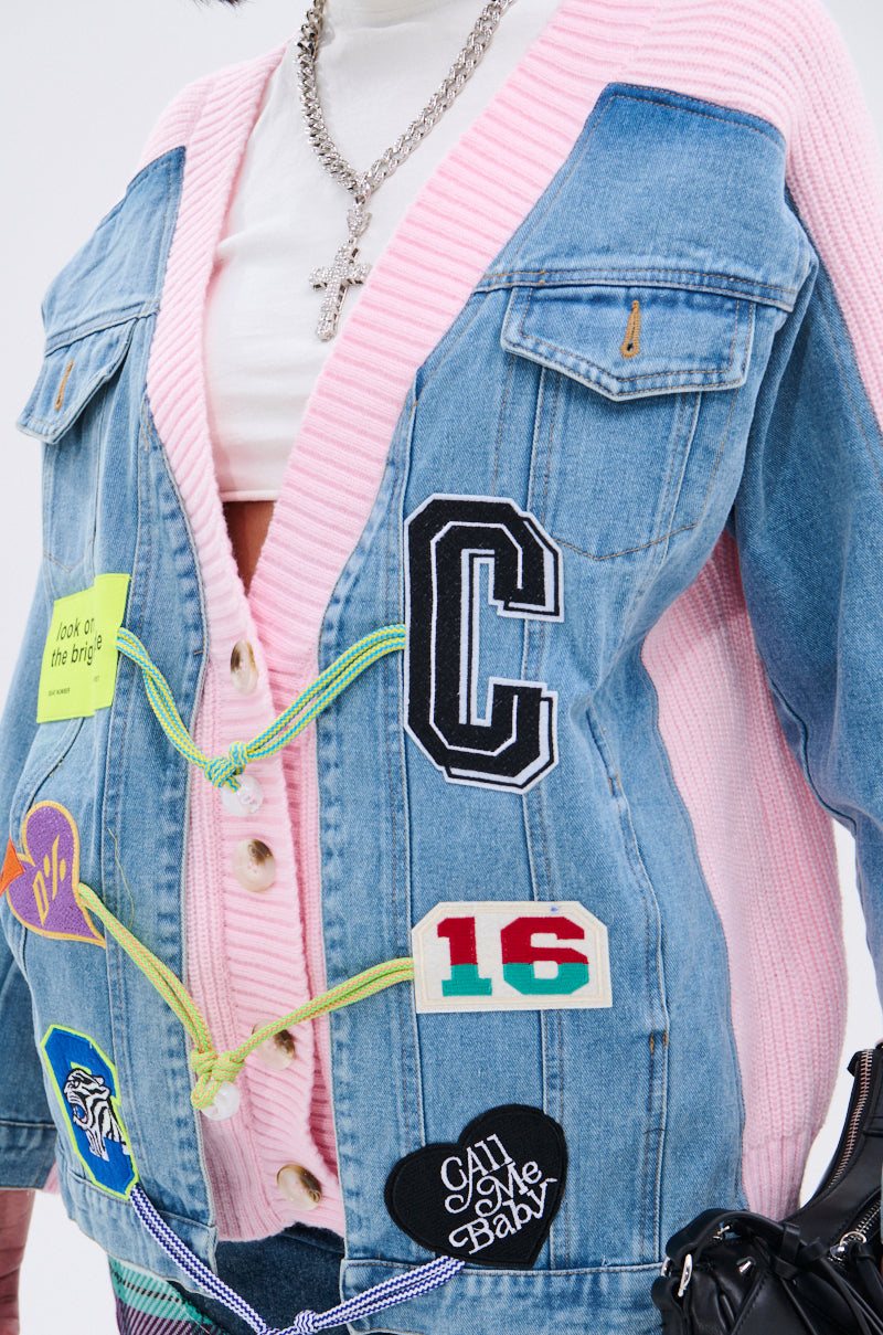 A person wears the SURGICAL SPLICED CARDIGAN in pink knit under a blue denim patch jacket with "C," "16," and colorful designs, accessorized with a silver chain necklace featuring a cross pendant. 12