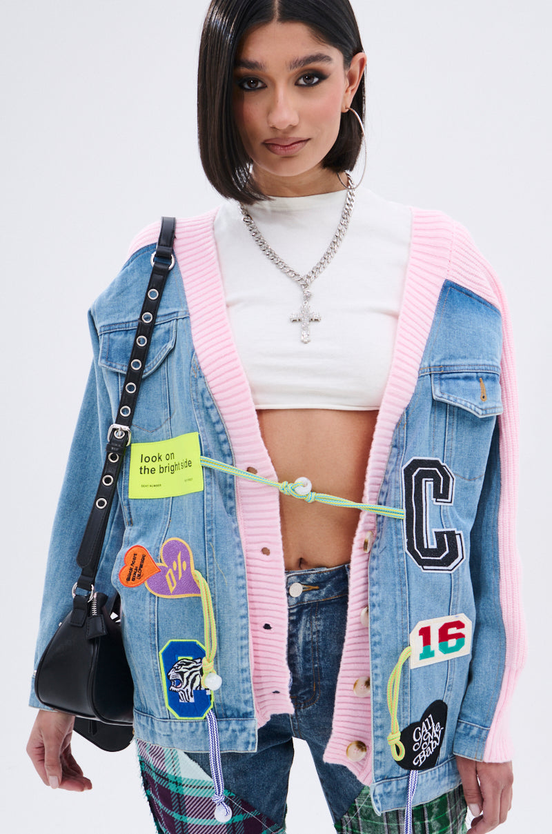 A woman with straight dark hair wears the SURGICAL SPLICED CARDIGAN, a blue denim hybrid sweater with colorful patches, pink trim, and green cords over a white crop top. She accessorizes with a black purse and silver jewelry. 1