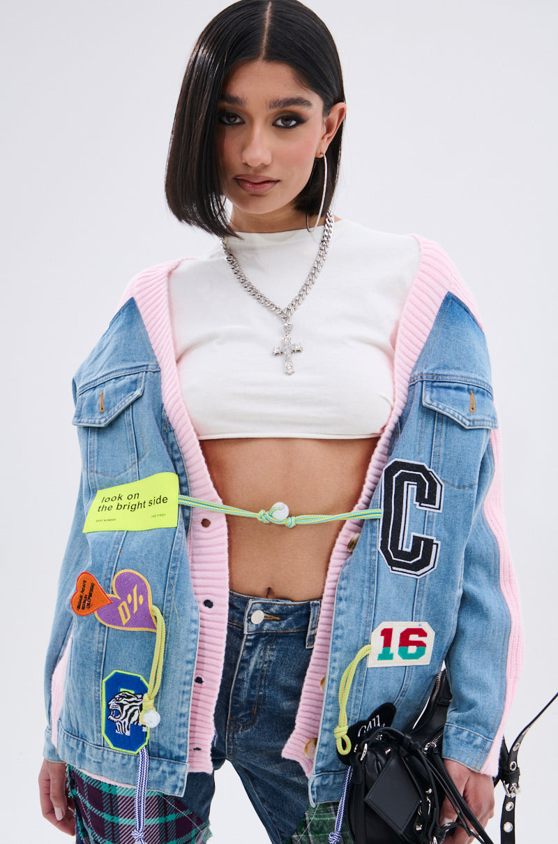 A young woman with straight dark hair wears a white crop top layered with the SURGICAL SPLICED CARDIGAN—a denim and pink hybrid sweater with colorful patches and messages—plus hoop earrings, a cross necklace, and a black bag. 2