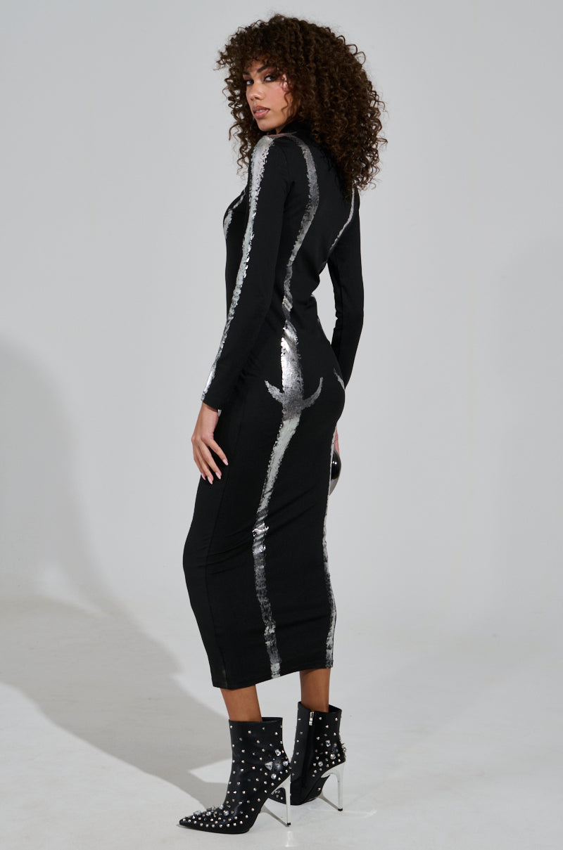 A woman with curly hair models the UNSEEN BITS ABSTRACT KNIT MIDI DRESS, a black ankle-length piece showcasing silver stripes and metallic detailing. She pairs it with black studded ankle boots and poses with side and back views against a plain background.