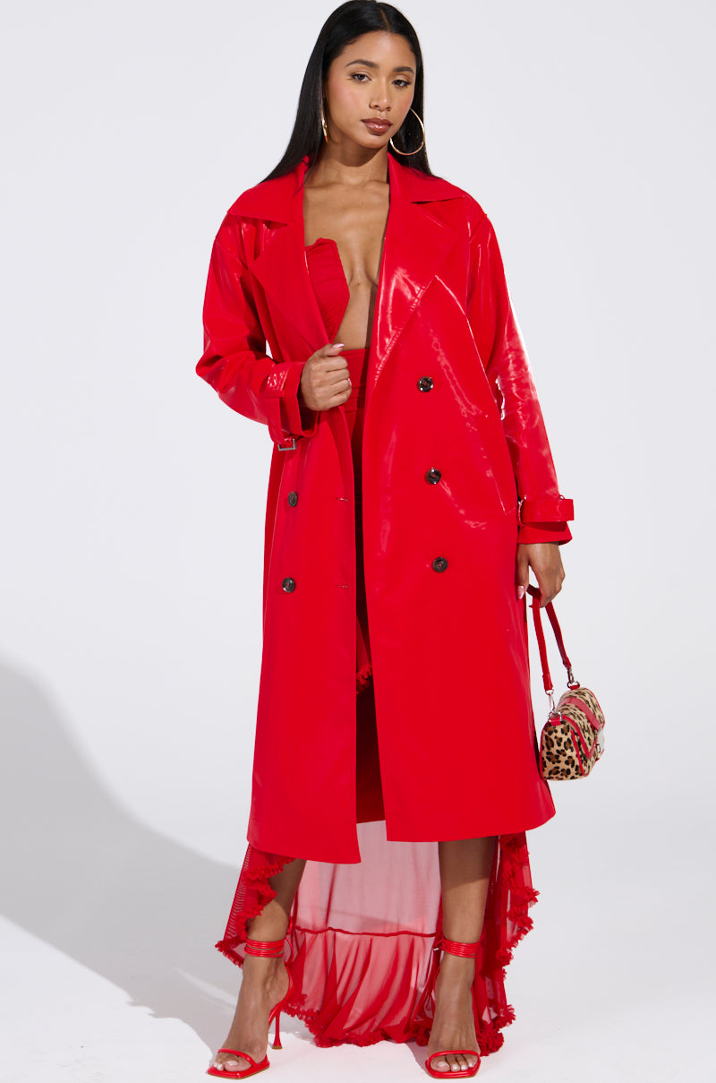 A woman stuns in the Spicy Sweet Liquid Velvet Trench, featuring a longline silhouette and a shiny red finish. Paired with matching high-heeled sandals and a small leopard print handbag, her open coat reveals a hint of her red dress beneath as she poses against a plain white background.