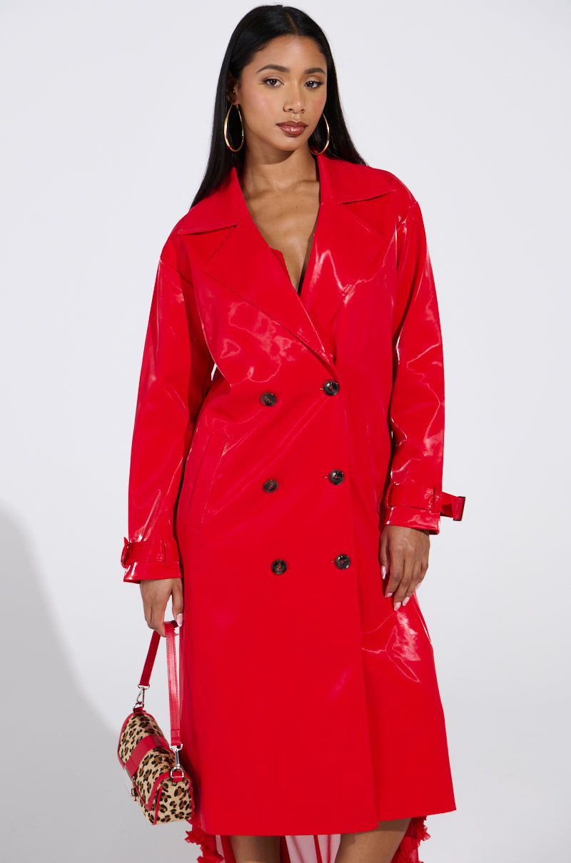 Against a plain backdrop, a person wears the vibrant SPICY SWEET LIQUID VELVET TRENCH with buttons and belt forming a longline silhouette. They hold a small leopard print handbag, flaunt long hair, and sport large hoop earrings.