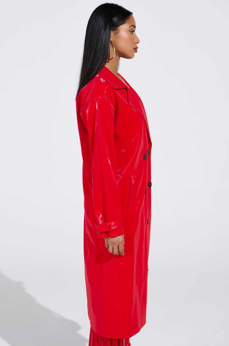 A person with long black hair is in profile, flaunting a shiny red SPICY SWEET LIQUID VELVET TRENCH. The coat's buttons and collar enhance its striking design against a plain white backdrop.