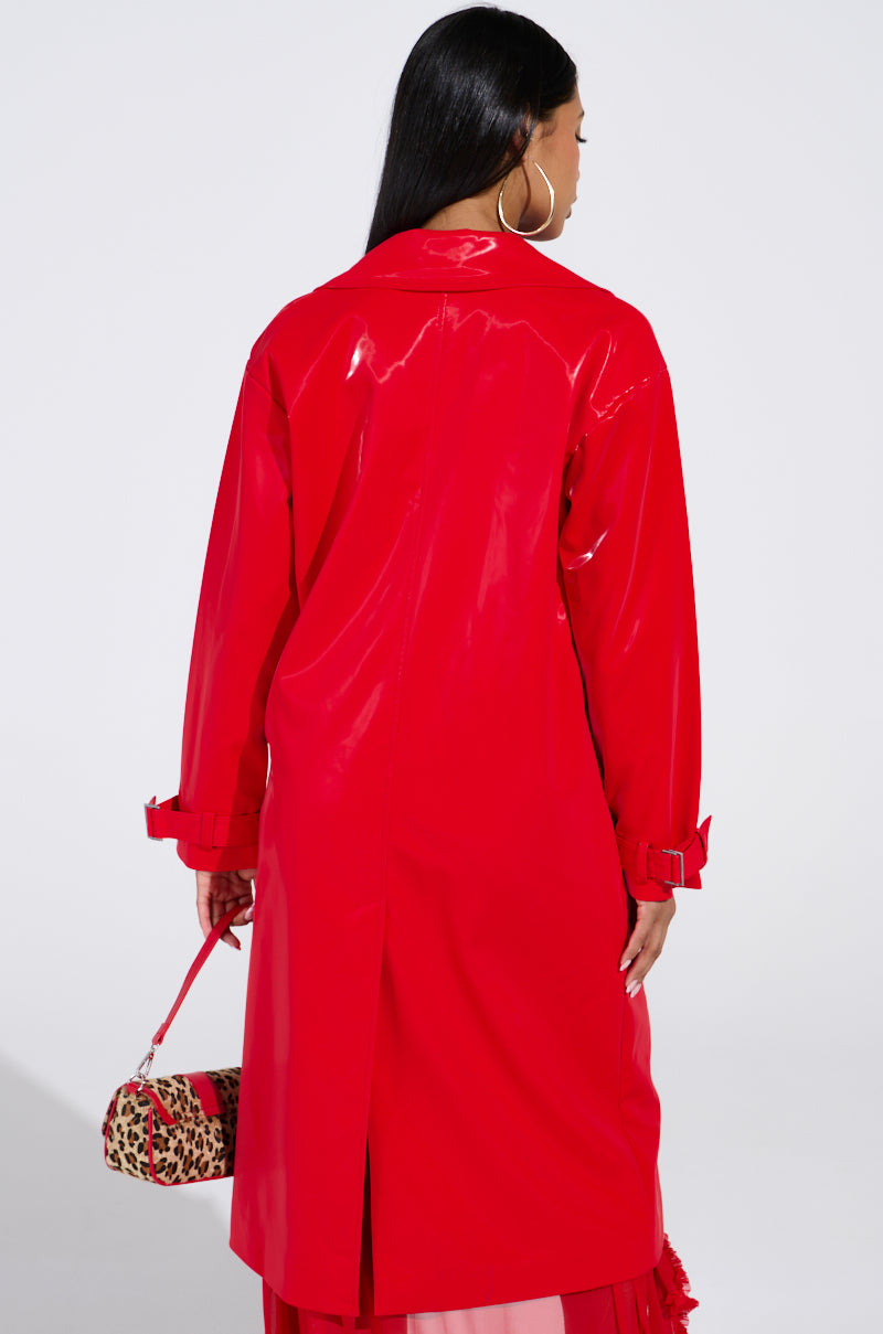 A person with long dark hair, facing away, wears the SPICY SWEET LIQUID VELVET TRENCH with a shiny red sheen and longline silhouette, holding a small leopard print bag against a plain, neutral background.