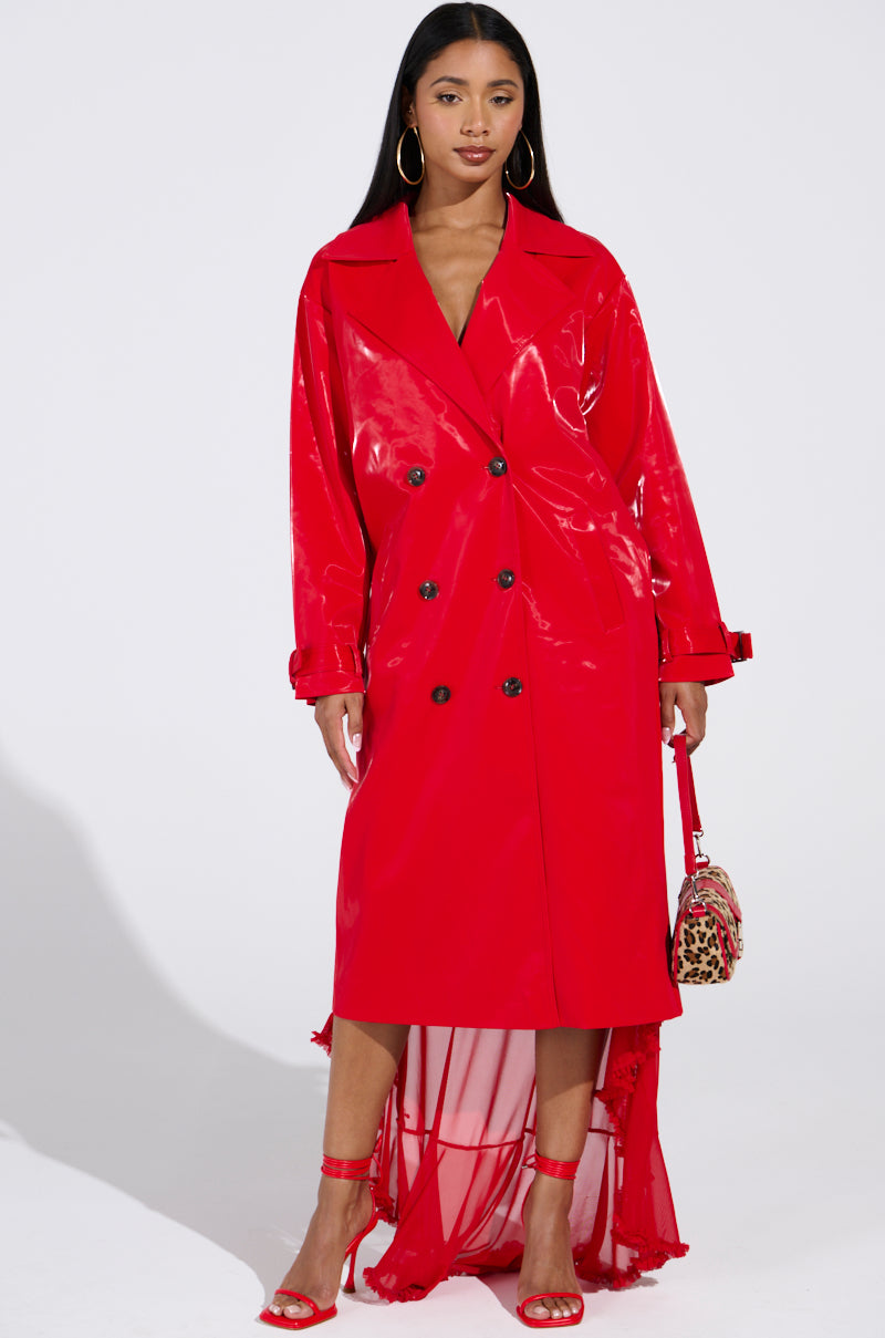 A woman exudes confidence in a SPICY SWEET LIQUID VELVET TRENCH with its shiny red longline silhouette. She accessorizes with a leopard print bag, red heeled sandals, loose hair, and hoop earrings against a plain background for a striking effect.