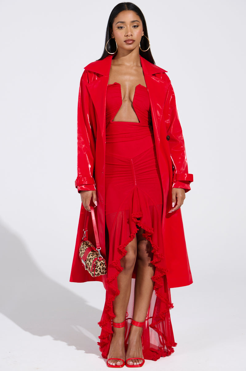 A confident woman stands against a plain background in a vibrant red outfit, featuring the Spicy Sweet Liquid Velvet Trench over a strapless dress with a ruffled high-low hem. She accessorizes with a leopard print handbag, large hoop earrings, and striking red heels.