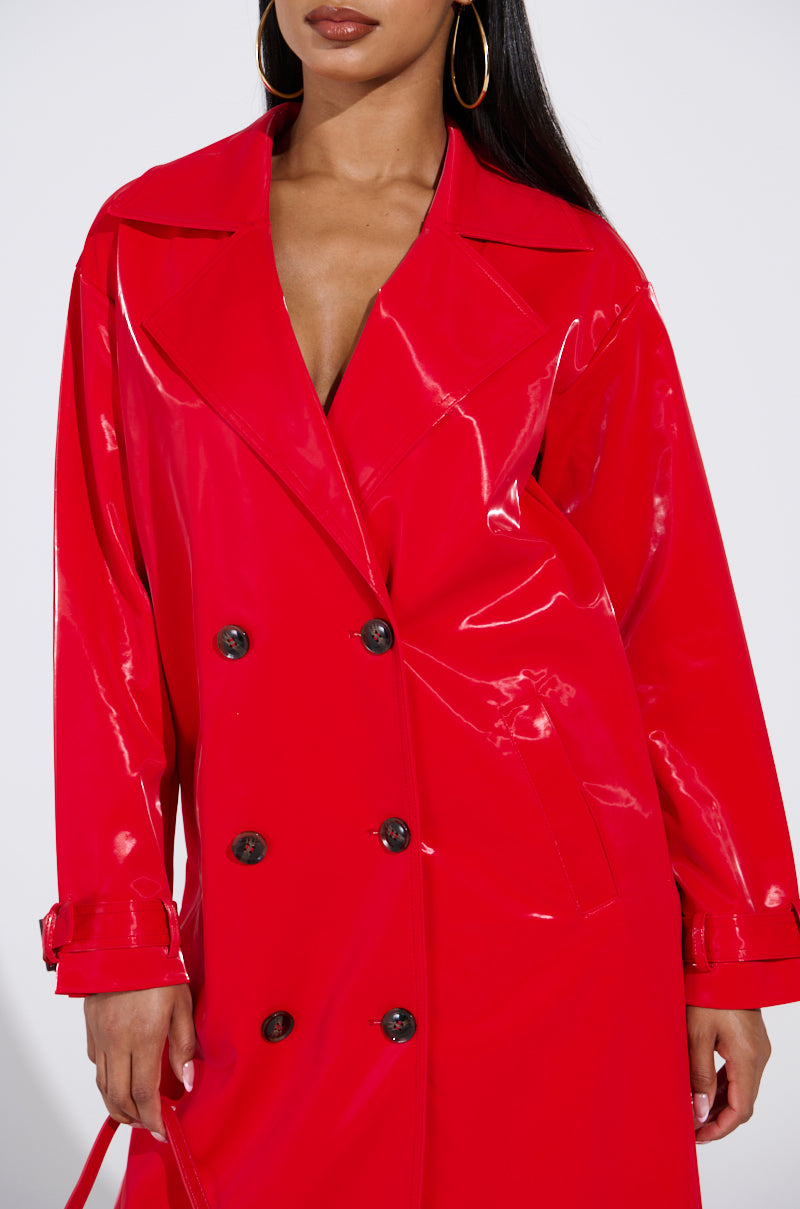 A person models a shiny red Spicy Sweet Liquid Velvet Trench featuring large buttons and belted cuffs, paired with gold hoop earrings. The photo highlights the trench's longline silhouette against a minimalist backdrop, focusing on the upper body.