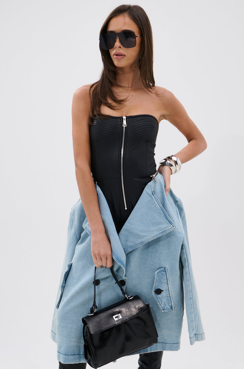 A woman poses against a white background, wearing a strapless black zip-up top, oversized sunglasses, and a large bracelet. She drapes the BUSINESS IN THE FRONT DENIM BLAZER MIDI DRESS over her arms and holds a black handbag.