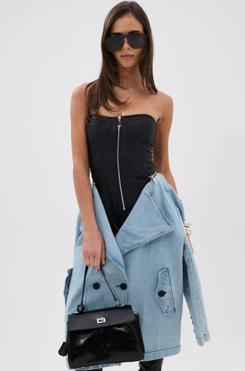 A woman in oversized black sunglasses, a strapless faux leather bodysuit, and the BUSINESS IN THE FRONT DENIM BLAZER MIDI DRESS draped over her arms holds a small black handbag against a plain white background.