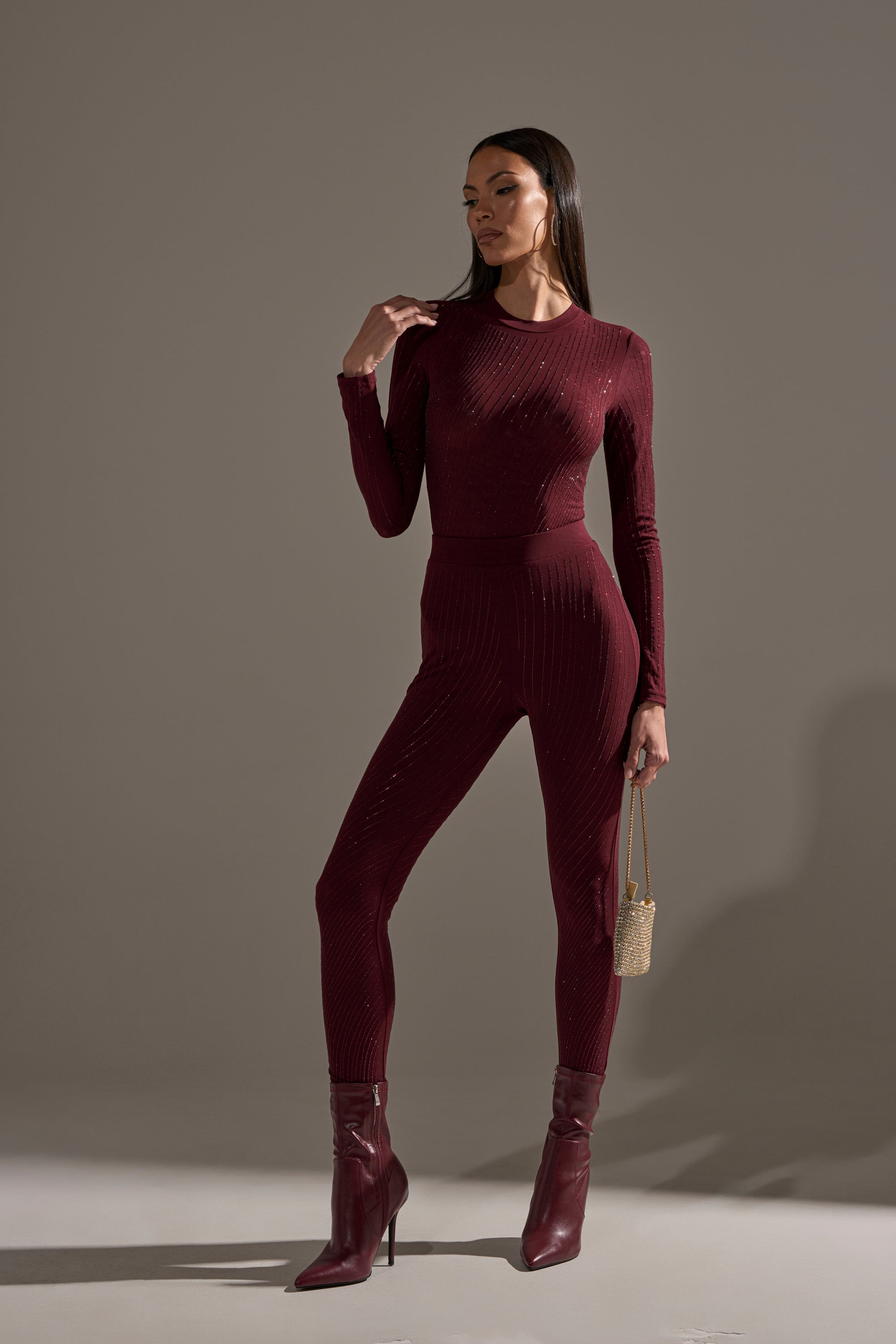 A woman stands confidently in TWINKLING LIGHTS RHINESTONE MESH LEGGINGS IN BURGUNDY, paired with a matching bodysuit, high-heeled boots, and a gold handbag, posing against a plain neutral background.