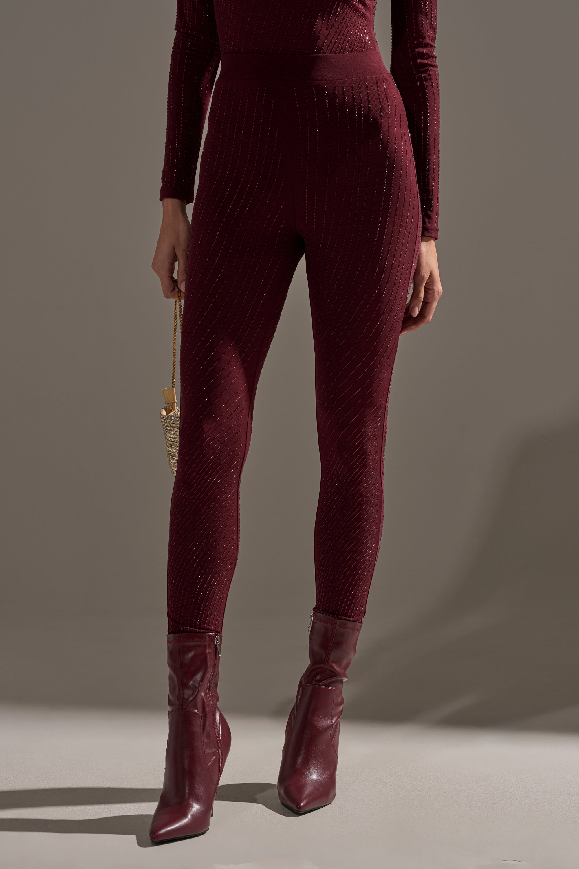 A person stands against a plain background wearing the TWINKLING LIGHTS RHINESTONE MESH LEGGINGS IN BURGUNDY, paired with a burgundy long-sleeve top, matching ankle boots, and holding a small gold purse.