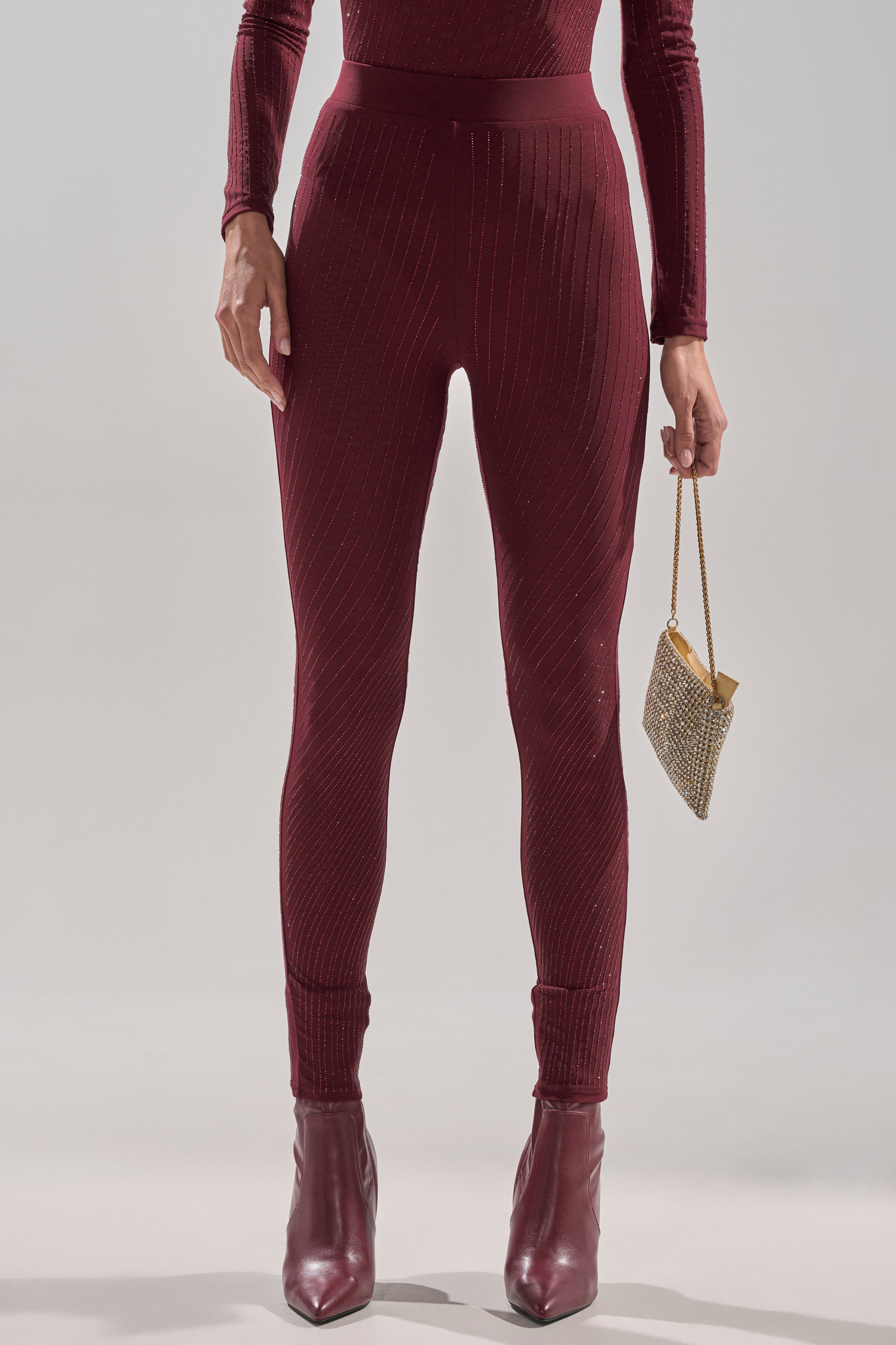 A person wears TWINKLING LIGHTS RHINESTONE MESH LEGGINGS IN BURGUNDY with a matching long-sleeve top, ankle boots, and holds a small gold purse in their right hand. The background is plain and light-colored.