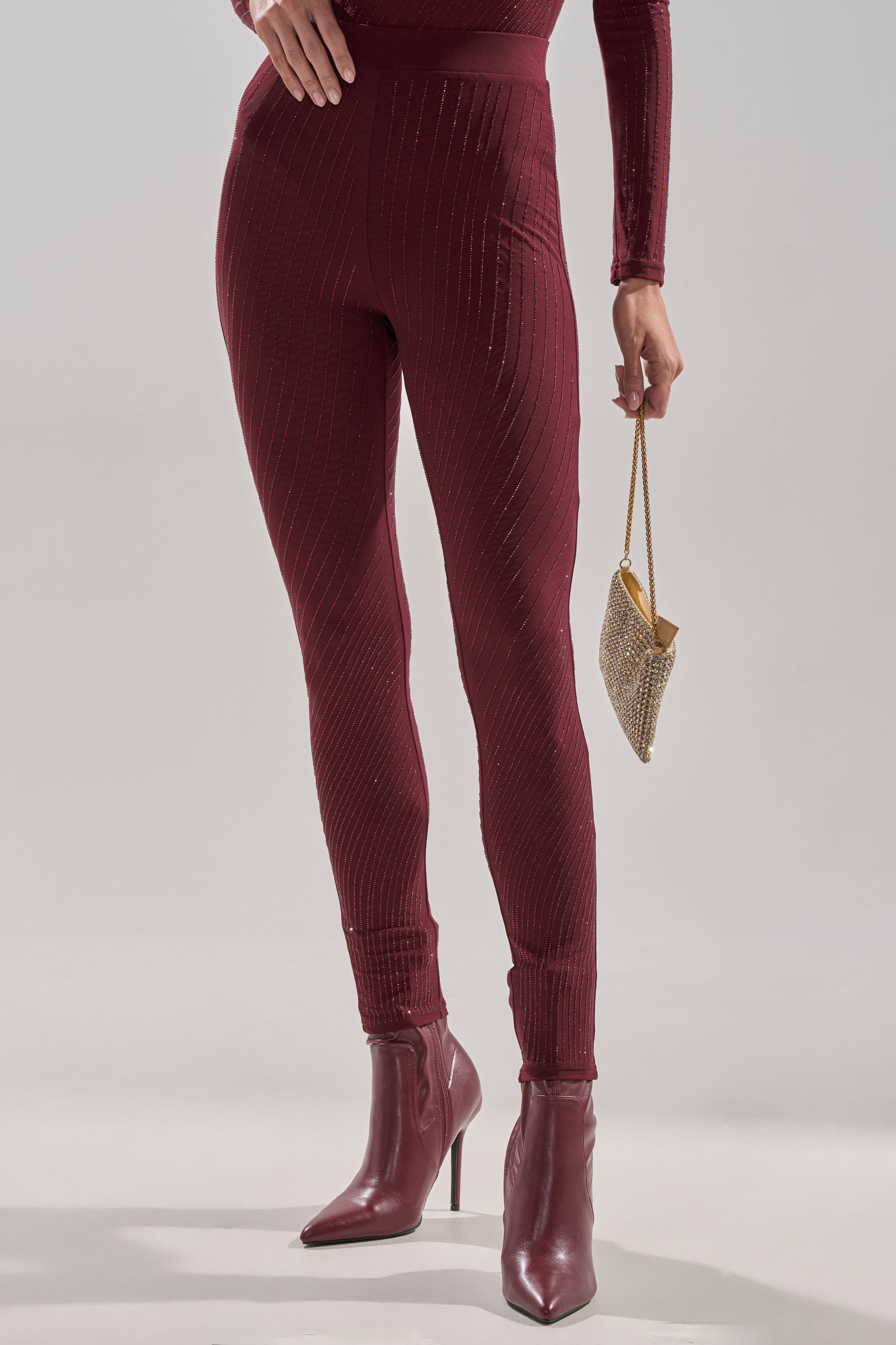 A person wearing TWINKLING LIGHTS RHINESTONE MESH LEGGINGS IN BURGUNDY with subtle vertical stripes, matching high-heeled ankle boots, and holding a small gold clutch against a plain light gray background.