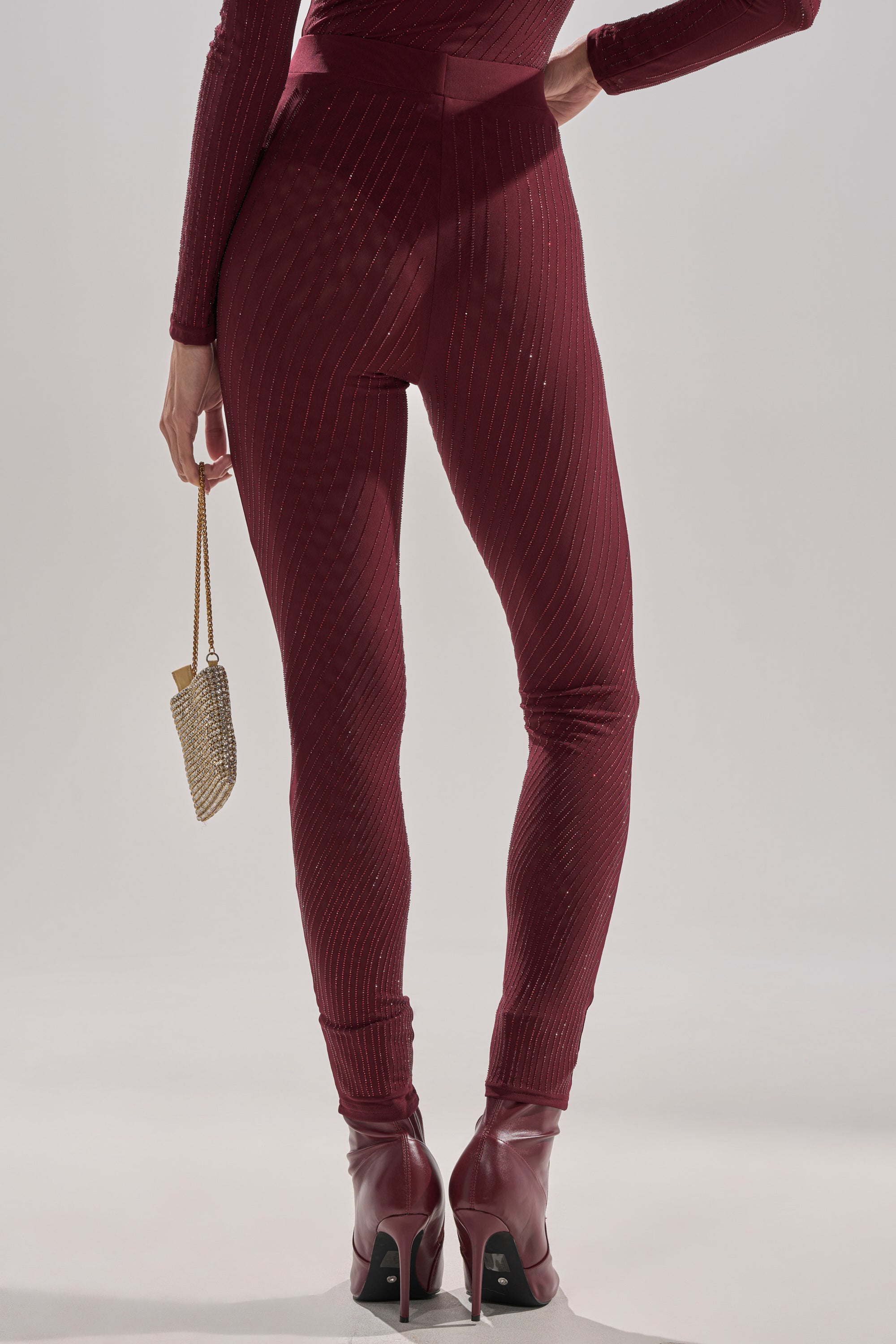 A person seen from behind wears the Twinkling Lights Rhinestone Mesh Leggings in Burgundy with a fitted, dark red bodysuit and heeled ankle boots, holding a small gold chain purse behind their back.