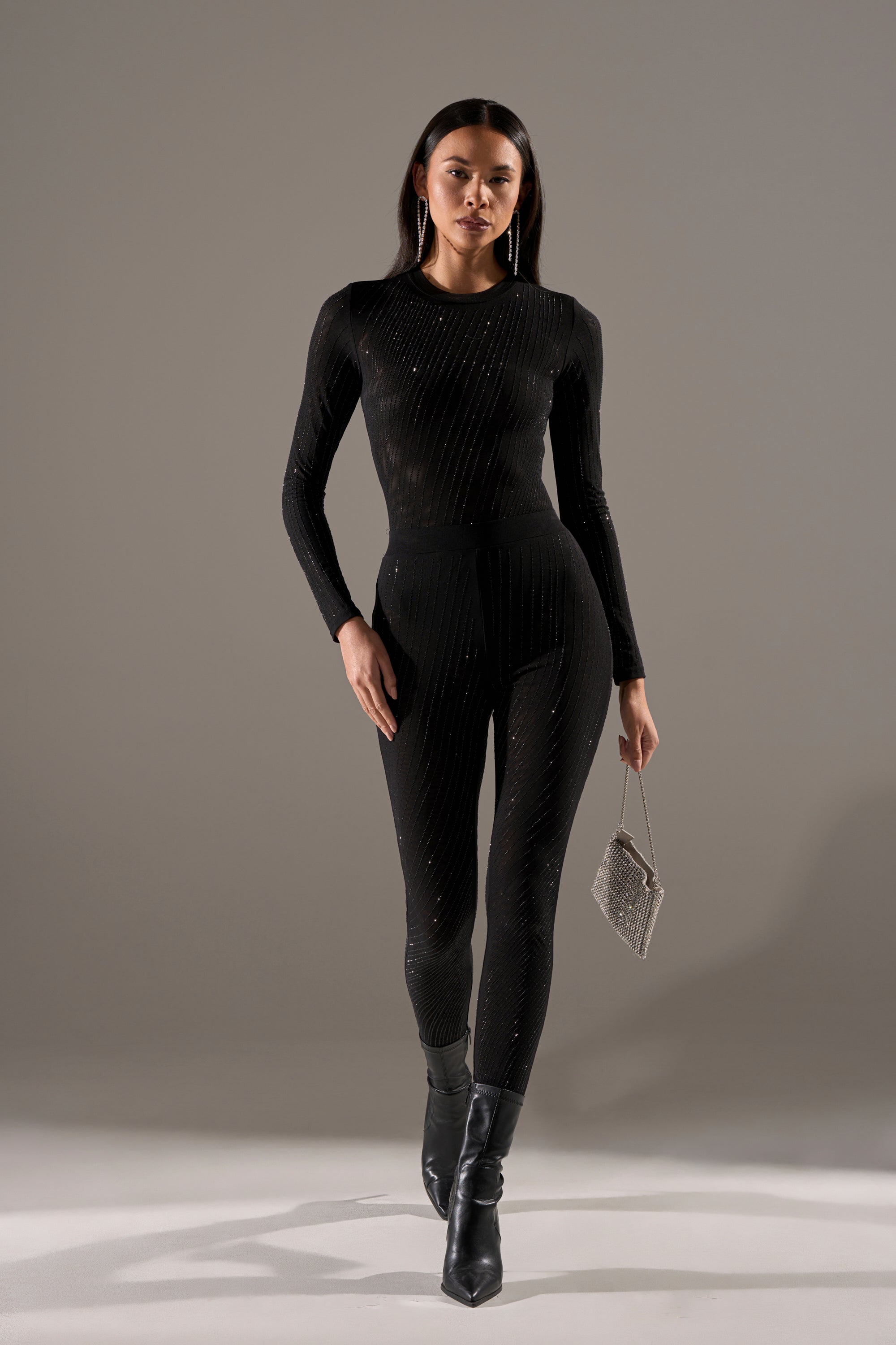 A woman stands confidently in a fitted, long-sleeve black jumpsuit with subtle sparkles, wearing TWINKLING LIGHTS RHINESTONE MESH LEGGINGS IN BLACK, a small silver clutch, and black ankle boots against a plain gray background.