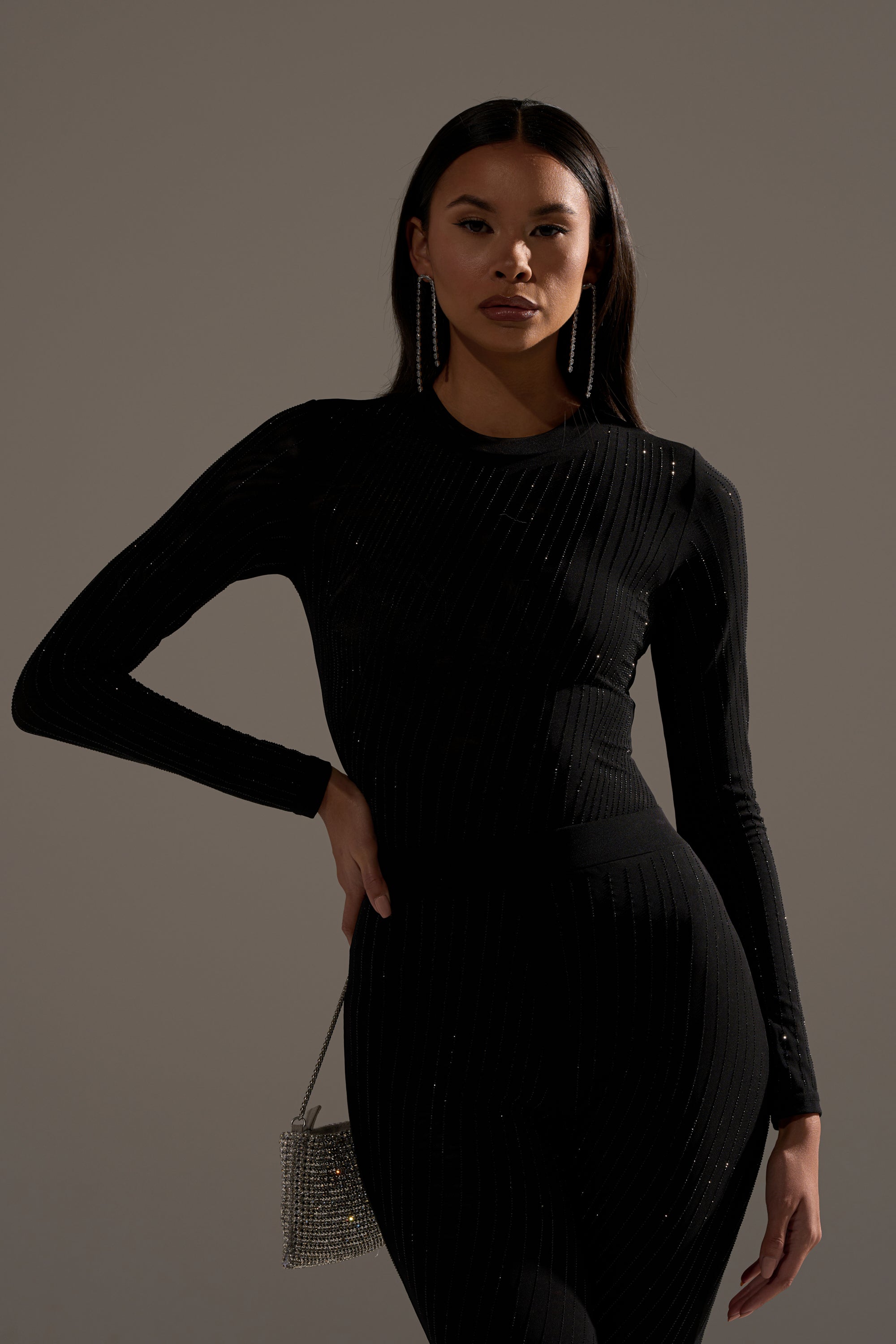 A woman confidently poses in the TWINKLING LIGHTS RHINESTONE LONG SLEEVE MESH BODYSUIT IN BLACK, featuring subtle sparkles. She accessorizes with dangling earrings and a glittery purse, standing against a plain background.