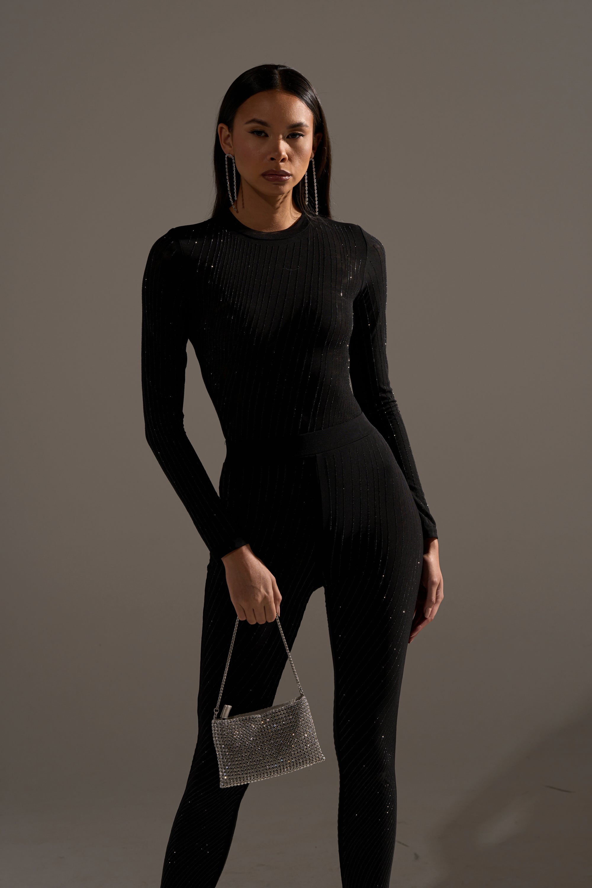 A woman stands against a plain background wearing the TWINKLING LIGHTS RHINESTONE LONG SLEEVE MESH BODYSUIT IN BLACK with matching pants. She holds a silver beaded handbag, wears hoop earrings, and has a neutral expression.