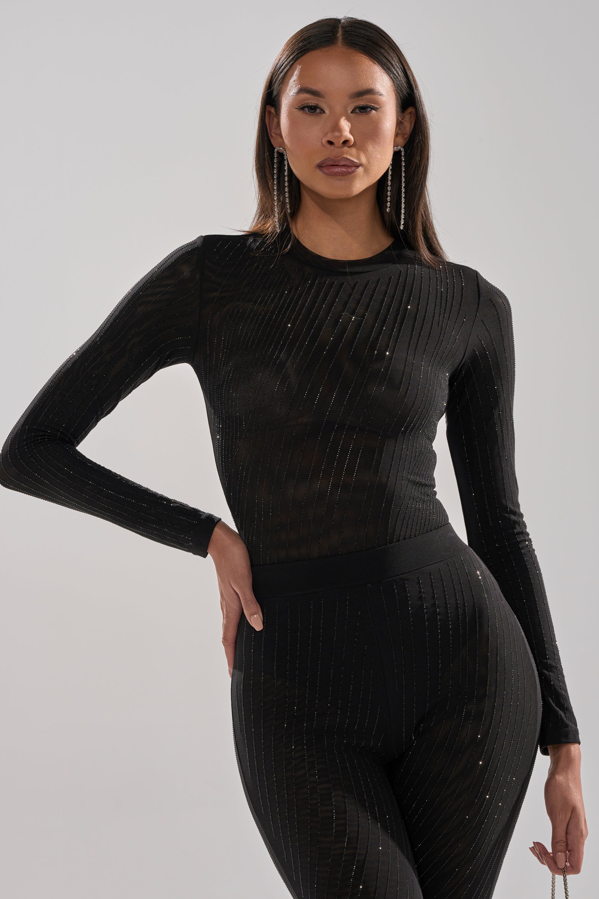 A woman poses confidently in the TWINKLING LIGHTS RHINESTONE LONG SLEEVE MESH BODYSUIT IN BLACK, featuring a fitted silhouette and subtle vertical sparkly stripes, against a plain light background.